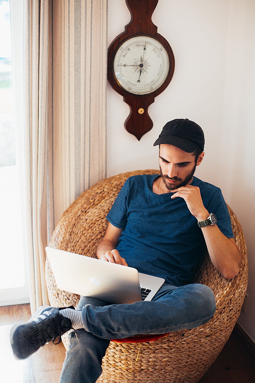 man in blue top sitting on wicker chair while using laptop